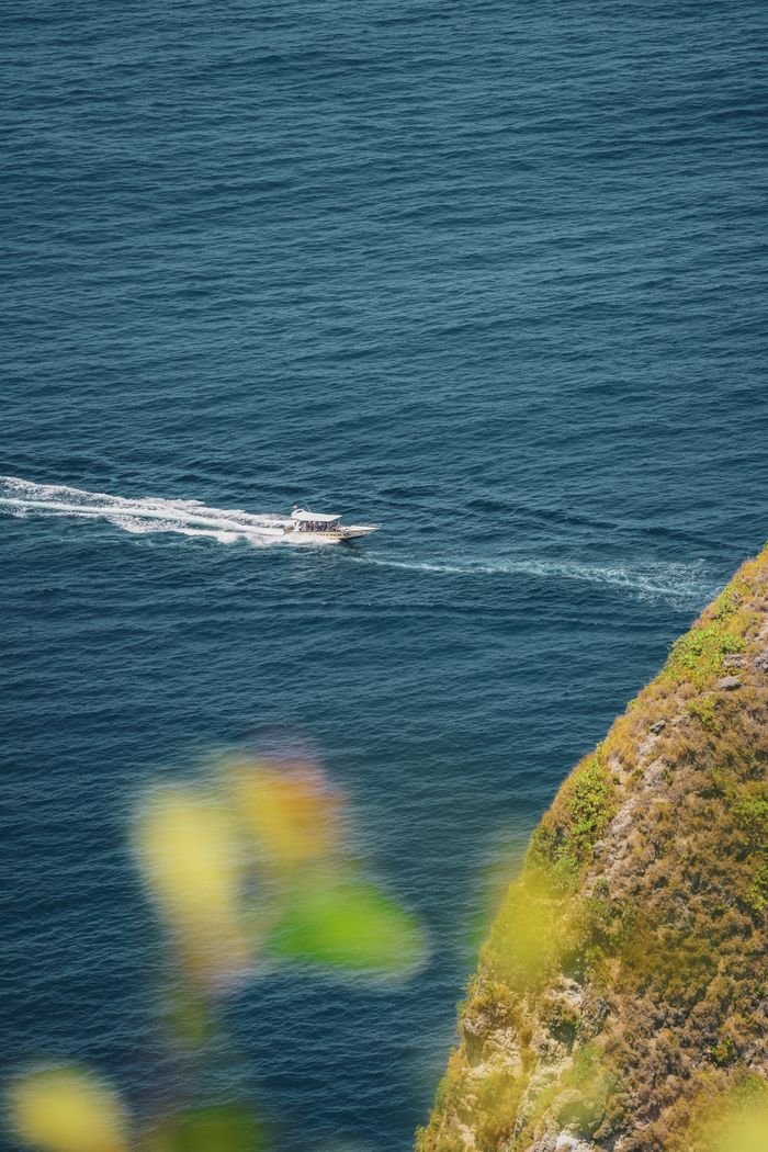 A boat glides through the blue ocean near Bali's lush cliffs, offering a beautiful coastal view.